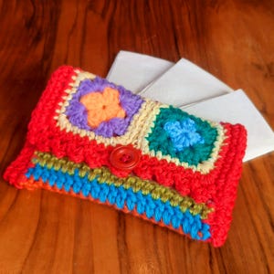 Puede incluir: Cartera de crochet hecha a mano en colores vibrantes. La cartera presenta una base roja con patrones cuadrados coloridos en morado, naranja, verde y azul. Un botón rojo asegura la solapa. Un accesorio único y artesanal.