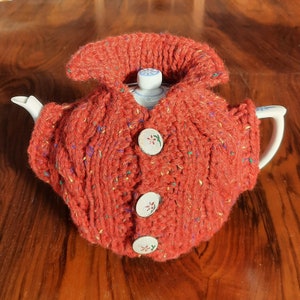 Könnte beinhalten: Eine rote gestrickte Teekannenhülle mit weißen Knöpfen und floralen Akzenten. Die Hülle ist aus einem dicken Garn gefertigt und hat ein Zopfmuster.
