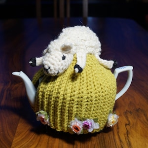 Puede incluir: Una tetera de cerámica blanca con un cubrete de té amarillo tejido. Un cordero blanco de ganchillo con pezuñas negras descansa encima. Pequeños adornos florales adornan la base del cubrete. La tetera está sobre una superficie de madera.