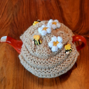 Peut inclure: Un chauffe-thé en tricot beige avec des fleurs blanches en crochet et des abeilles jaunes et noires.
