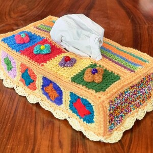 Può includere: Una copertura rettangolare per scatola di fazzoletti realizzata con quadrati all'uncinetto multicolori nelle tonalità del blu, rosso, giallo, verde e viola. La parte superiore presenta piccoli fiori all'uncinetto con perline e un bordo in pizzo color crema.