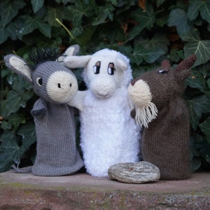 Peut inclure: Trois marionnettes à doigts en tricot, un âne gris, un mouton blanc et une chèvre brune, sont assises sur une surface en pierre. Les marionnettes regardent toutes le spectateur.