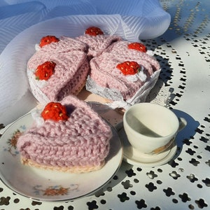 Könnte beinhalten: Drei gehäkelte rosa Erdbeerkuchen mit weißer Spitzenborte. Die Kuchen stehen auf einer weißen Oberfläche mit einer weißen Teetasse und Untertasse.