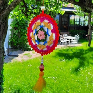 Könnte beinhalten: Ein bunter gehäkelter Traumfänger mit einem Blumenmuster in Pink, Grün, Blau und Gelb. Der Traumfänger hängt an einer weißen Perle und hat eine lange gelbe Quaste.