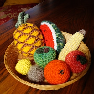 Op de afbeelding: Een rieten mand gevuld met gehaakte vruchten, waaronder een ananas, een watermeloen, een banaan, een groene appel, een sinaasappel en een rode appel.