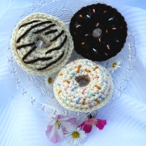 Op de afbeelding: Drie gehaakte donuts op een glazen bord. Eén donut is wit met bruine strepen, één is zwart met roze en witte sprinkles, en één is wit met oranje en blauwe sprinkles.