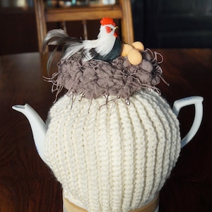 Könnte beinhalten: Weiße gehäkelte Teekannen-Cozy mit einem braunen Nest und einer weißen Hahnfigur oben drauf. Die Cozy befindet sich auf einer weißen Teekanne mit blau-weißem Design.