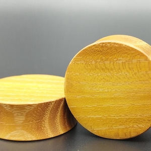 May include: Two wooden ear plugs with a light brown, golden hue. The plugs are cylindrical with a flared edge. The wood grain is visible, adding texture to the smooth surface. The plugs are designed for stretched ear piercings.
