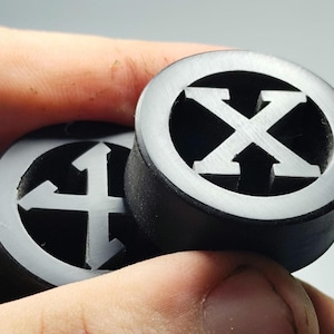 Peut inclure: Deux bouchons d'oreille noirs avec un motif circulaire et un logo blanc en forme de X. Les bouchons sont tenus dans une main, mettant en valeur le design détaillé et le contraste entre les éléments noirs et blancs.