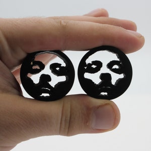 Könnte beinhalten: Zwei schwarze, kreisförmige Ohrstöpsel mit einem Gesichtsdesign. Das Gesicht ist aus dem schwarzen Material ausgeschnitten, wodurch ein negativer Raum entsteht, der den Hintergrund freilegt. Die Ohrstöpsel werden vor einem weißen Hintergrund gehalten.