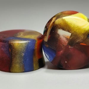 May include: Two colorful ear plugs. One is cylindrical and the other is rounded. Both are made of a translucent material with red, yellow, blue, and gold colors. The plugs have a marbled appearance.