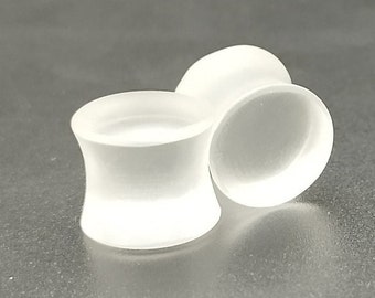 May include: Two white, frosted glass ear gauges. The gauges have a flared, hourglass shape with a flat, circular face. The gauges are smooth and opaque, with a soft, diffused appearance. The gauges are designed for stretched ear piercings.