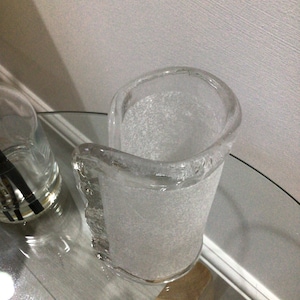 May include: A clear glass vase with a textured surface. The vase has a unique, curved shape and a frosted finish.