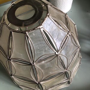 May include: A dome-shaped lampshade constructed from translucent, off-white shell panels held together by a dark metal framework. The structure features a repeating geometric pattern of intersecting petal shapes and a circular metal opening at the top.