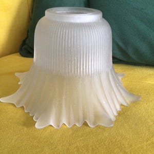 May include: A frosted glass lampshade with a flared, scalloped edge. The shade has vertical ribbed detailing and a soft, diffused glow. The lampshade is off-white and sits on a yellow surface.