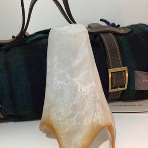 May include: A white, textured, glass lampshade with a brown, irregular base. The lampshade is sitting on top of a green and black plaid blanket with a brown leather strap.