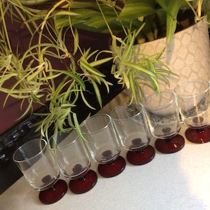 May include: Six clear glass tumblers with a red base are arranged in a row. The glasses are empty and angled slightly. A white surface and a plant in a decorative pot are in the background.