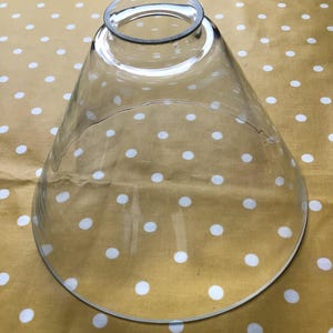 May include: A clear glass light shade with a conical shape, featuring a wide circular base and a smaller circular opening at the top. The glass is transparent and smooth, designed to fit onto a light fixture, showcasing a simple, functional form.