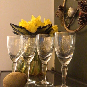 May include: Four clear glass wine glasses with a starburst pattern sit on a dark surface. A vase with yellow daffodils sits behind the glasses.