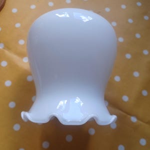 May include: A white glass lampshade with a frilled edge. The lampshade has a rounded top and a flared, scalloped bottom. The surface is smooth and reflective, with highlights from the light source. The lampshade is designed to diffuse light.