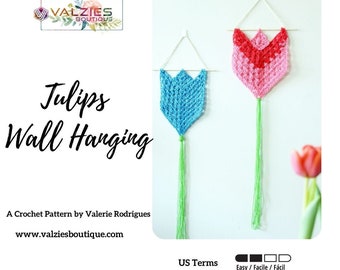 Op de afbeelding: Twee gehaakte tulpen wanddecoraties, een blauwe en een roze, met groene garenkwastjes. De decoraties worden aan een witte muur gehangen met een houten stokje bovenaan. De tekst "US Terms Easy / Facile / Fácil" staat onder de decoraties.