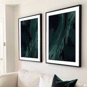 Puede incluir: Dos pinturas abstractas enmarcadas con marcos negros y un passe-partout blanco. Las pinturas son ambas verdes y negras con pinceladas abstractas. Las pinturas están colgadas en una pared blanca sobre un sofá blanco con un cojín negro y verde.