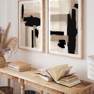 May include: Two framed abstract art prints featuring black and beige brushstrokes on a white background. The prints are hanging on a white wall above a wooden console table with books and a vase.