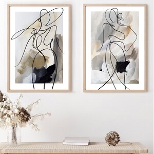 May include: Two framed abstract art prints with black line drawings of figures on a beige background with white and black paint splatters.