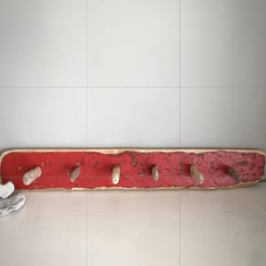 May include: A long, rectangular wooden wall rack featuring a distressed, weathered red paint finish. The piece includes six light-coloured, cylindrical wooden pegs protruding from the front surface for hanging items.