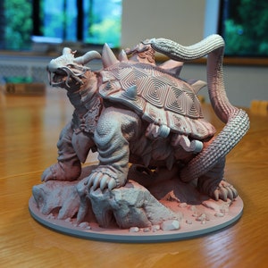 Könnte beinhalten: Detaillierte Skulptur einer Fantasy-Kreatur, ein schildkrötenartiges Tier mit Hörnern, Stacheln und einer Schlange, die sich darum windet. Die hellfarbene Skulptur steht auf einem runden Sockel mit filigranen Details.