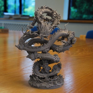 Könnte beinhalten: Eine graue, detaillierte Drachenskulptur mit einem eingerollten Schwanz und Flügeln, in einer dynamischen, gewundenen Position. Der Drache ist mit komplizierten Schuppen und einem grimmigen Ausdruck modelliert.