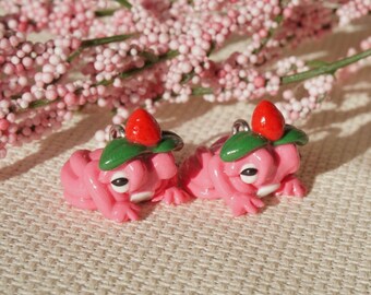 Può includere: Due figurine di rana rosa, ognuna ornata da una fragola rossa e foglie verdi sulla testa. Le rane hanno occhi neri, su uno sfondo di rami floreali rosa.