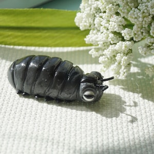 May include: A detailed, dark gray sculpture of a pill bug, also known as a roly-poly. The sculpture has a segmented body, small antennae, and realistic detailing. The background includes white flowers and a green leaf.