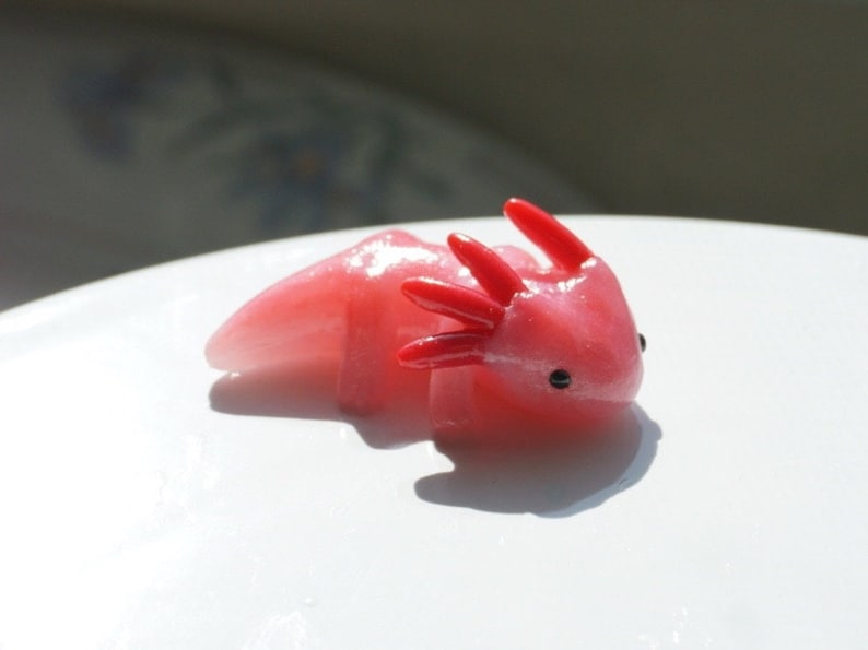 May include: A pink axolotl figurine with red gills and small black eyes. The figurine is resting on a white surface, with the light casting a shadow to the left. The axolotl is a salamander known for its unique appearance and ability to regenerate limbs.