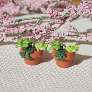 May include: A pair of miniature terracotta-colored flower pots, each containing a green plant and a small, light green frog. The frog is peeking out from the plant. The background features pink flowers.
