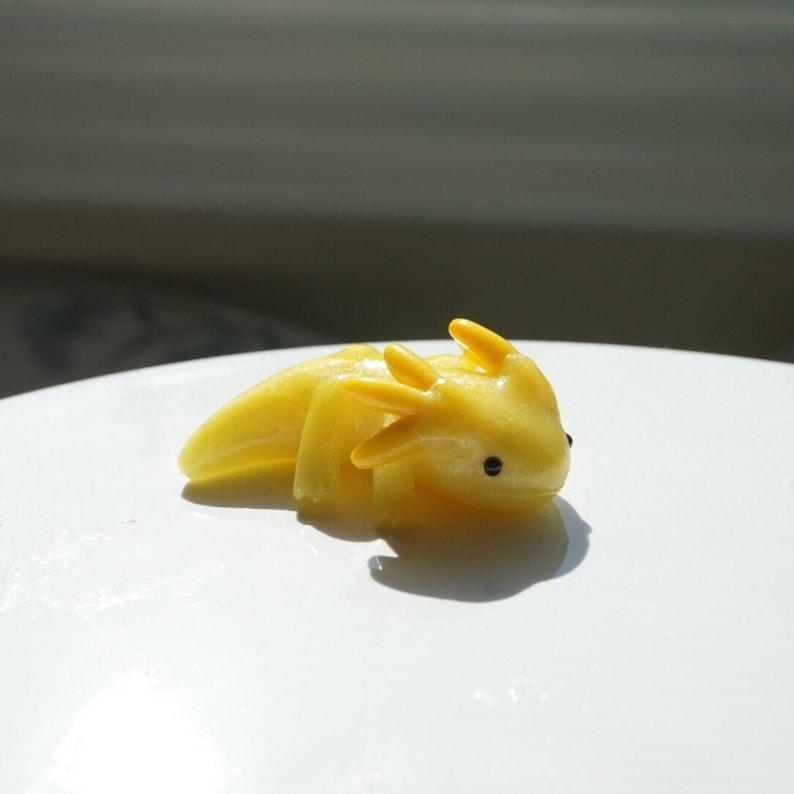 May include: A small, yellow axolotl figurine with four external gills and tiny black eyes. The figurine is resting on a white surface, and the lighting creates a shadow to the right of the axolotl.