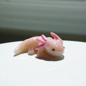 May include: A small, light pink axolotl figurine with iridescent glitter accents. The figurine has pink gills and small, dark eyes. The axolotl is lying down on a white surface, with sunlight in the background.