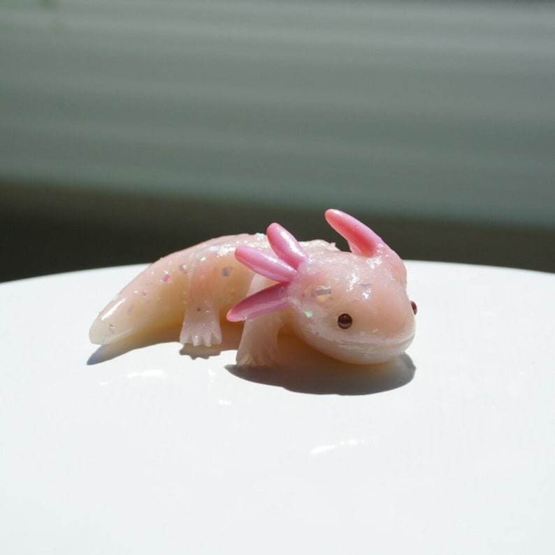 May include: A small, light pink axolotl figurine with iridescent glitter accents. The figurine has pink gills and small, dark eyes. The axolotl is lying down on a white surface, with sunlight in the background.