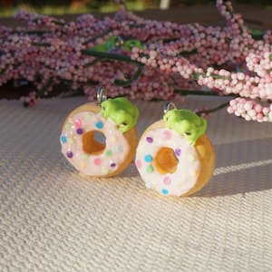 May include: Two donut-shaped charms with pink icing and colourful sprinkles. Each charm features a small green frog on top. The charms are made of polymer clay and have silver loops for hanging, suitable for jewellery.