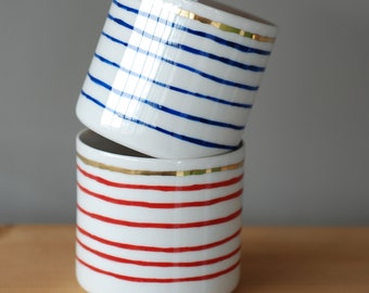 May include: Two white ceramic cups with gold trim and striped patterns. The top cup has blue stripes and the bottom cup has red stripes.