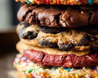 May include: A stack of six cookies, each with a different flavour and topping. The top cookie is covered in colourful sprinkles, the next is a chocolate cookie, followed by a cookie with a chocolate frosting, a red velvet cookie, a cookie with colourful sprinkles, and a plain cookie.