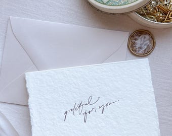 May include: A white rectangular card with deckled edges featuring the black handwritten text grateful for you. The card is positioned next to a light-colored envelope, a round metal tin with a vintage illustration on the lid, and a small wax seal.