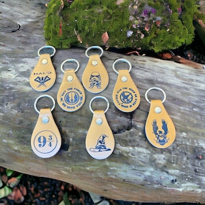 May include: A collection of tan leather keychains with silver key rings. Each keychain features a different design, including text and graphics from popular franchises like Halo, Star Wars, and Harry Potter. The keychains are arranged on a wooden surface.