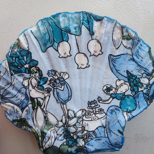 May include: A blue and white seashell with a painted scene of three fairies having tea. The fairies are wearing blue dresses and have wings. The scene is surrounded by flowers and leaves.