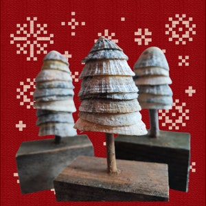 May include: Three small Christmas trees made of seashells on wooden bases. The trees are decorated with white and grey shells and are arranged on a red background with a snowflake pattern.