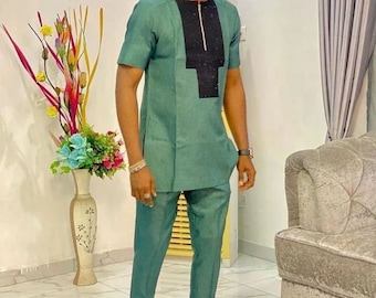 May include: A man wearing a green short-sleeved shirt with a black and silver zipper and a black panel with silver embellishments. He is also wearing matching green pants and black shoes.