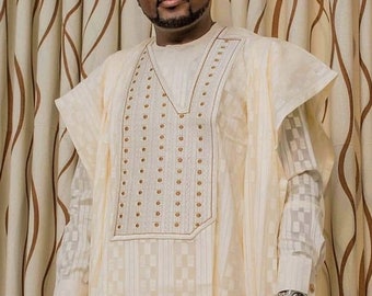 May include: A man wearing a white traditional garment with a checkered pattern and gold embroidery. He is wearing a black hat with a patterned design and sunglasses. He is also wearing a watch on his left wrist.