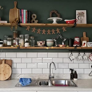 May include: A kitchen scene featuring two wooden shelves against a dark green wall above a white subway tile backsplash. The shelves hold various items including a wooden cutting board, books, glass jars, a gingerbread man garland, and mugs.