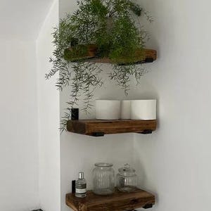 May include: Three wooden shelves with black metal brackets mounted on a white wall. The shelves are decorated with potted plants and glass jars.