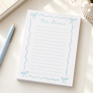 Puede incluir: Un bloc de notas rectangular blanco con un borde azul claro de líneas onduladas y lazos en cada esquina. El texto Mrs. Briceno está impreso en letra cursiva azul claro en la parte superior, seguido de líneas horizontales azul claro.